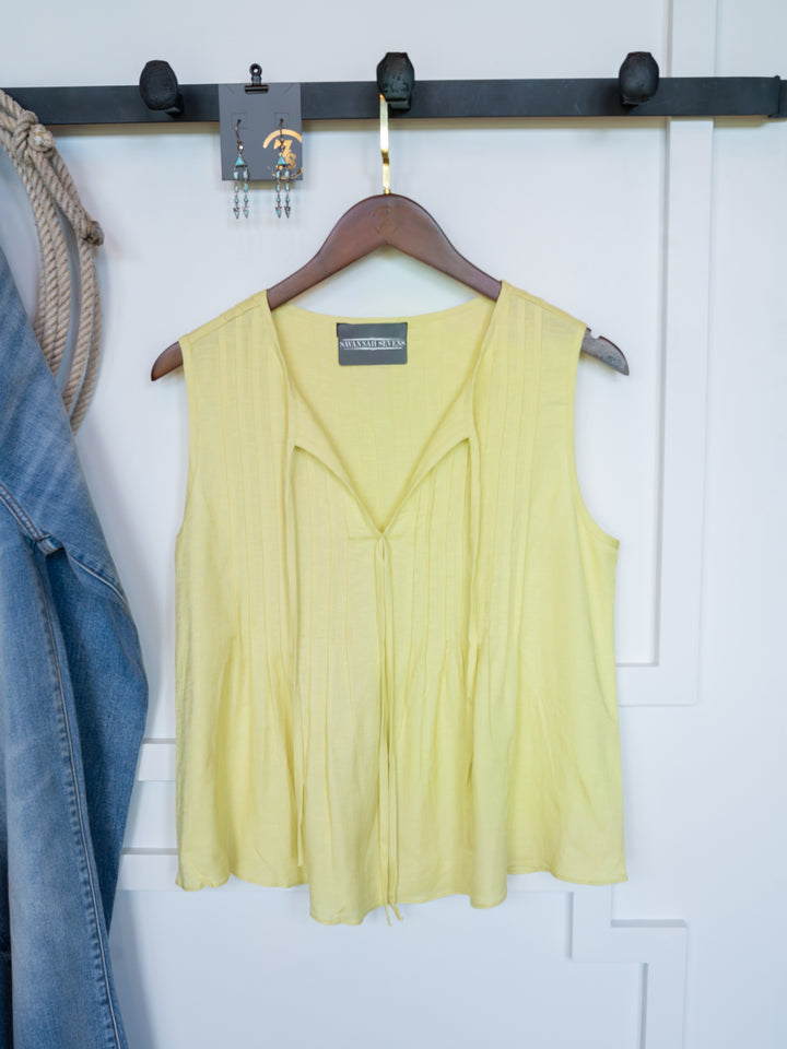 The Marlette by Savannah Sevens western life{&}style, a chartreuse pleat top with an adjustable tie neckline, hangs on a wooden hanger against a white wall. A denim jacket and earrings are also visible.