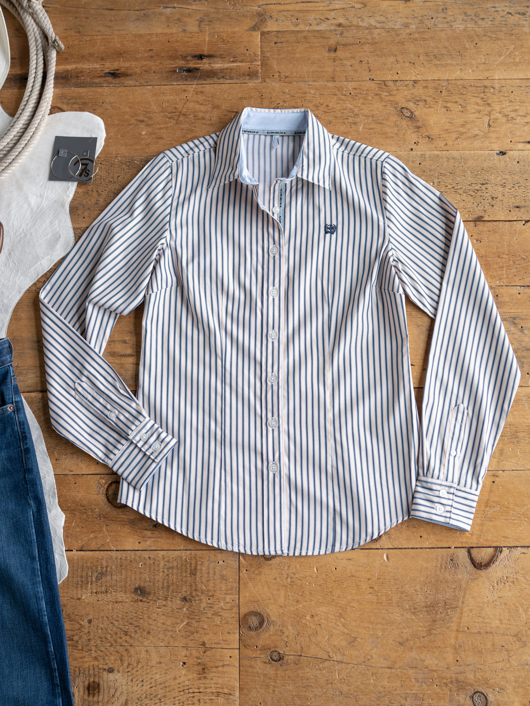 The Durant (S-XXL) by Savannah Sevens western life{&}style—a white women’s fitted shirt with blue vertical stripes—is neatly displayed on a wooden floor, with glimpses of a white tote bag and blue jeans at the edges.