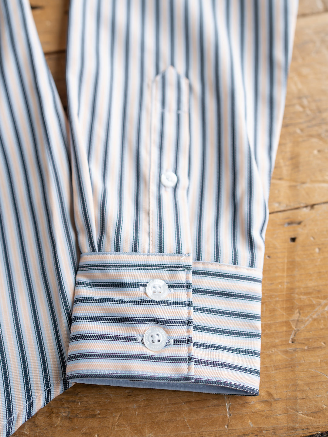 A close-up of The Durant women’s fitted shirt by Savannah Sevens western life{&}style, showing blue, white, and beige vertical stripes. The long sleeve button-up has three white buttons on the cuff and rests on a wooden surface.