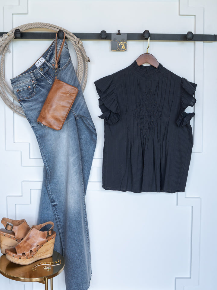 The Winnett by Savannah Sevens western life{&}style—a blue jean, midnight navy top, brown leather wristlet, and tan wedge sandal ensemble—is arranged neatly against a white wall with trim and a black hook rack.