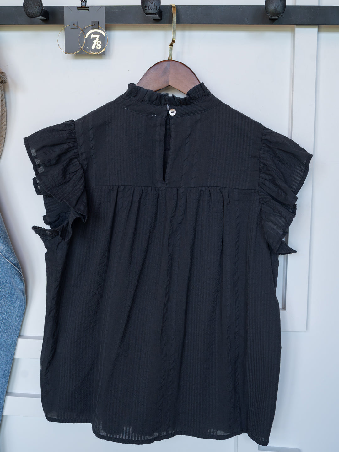 The Winnett by Savannah Sevens western life{&}style is a black cotton ruffle blouse with short ruffled sleeves, a high neckline, subtle texture, and a back button closure, displayed on a wooden hanger against a white background.