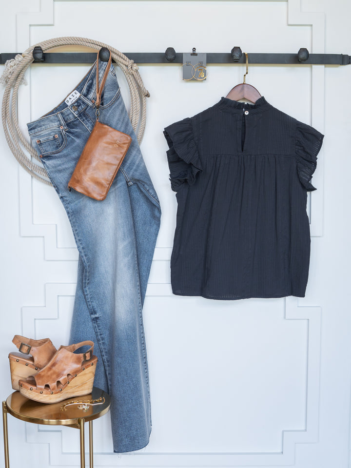 The Winnett by Savannah Sevens western life{&}style—a chic outfit with blue jeans, a cotton ruffle blouse, tan leather wristlet, and tan wedge sandals—is displayed neatly against a white wall with molding and a black hook rack.