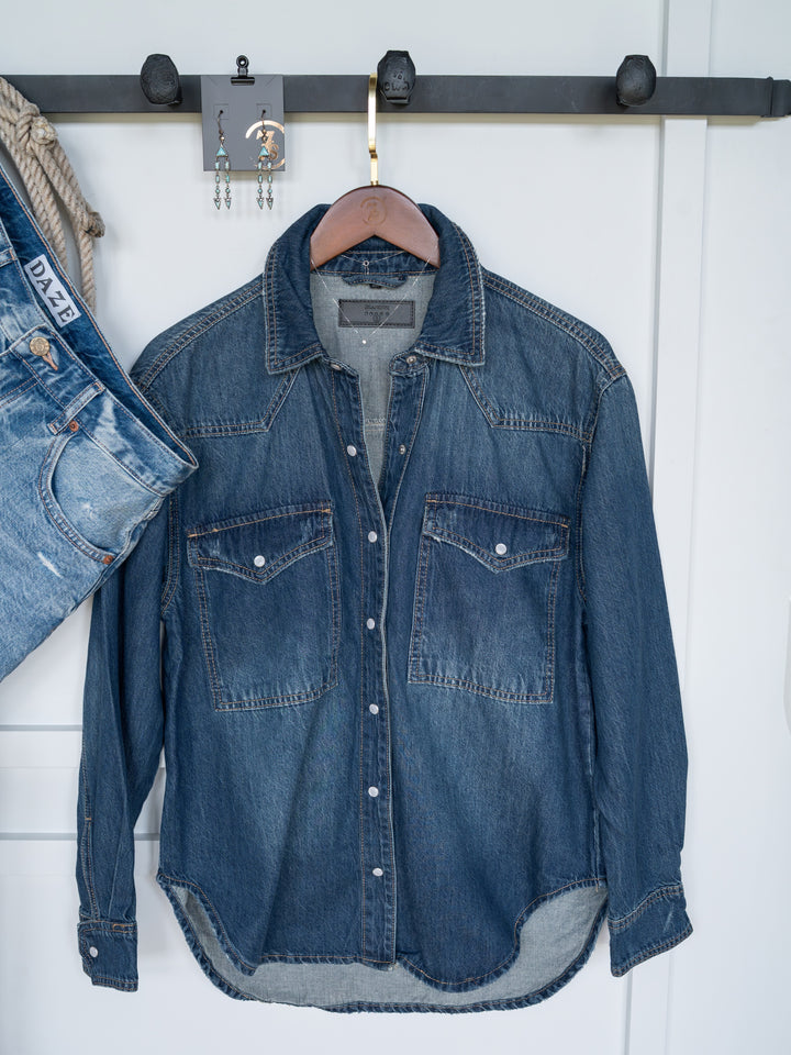 The Sorento by Savannah Sevens western life{&}style, a distressed denim shirt with a western yoke and snap buttons, hangs on a wooden hanger beside blue jeans and colorful earrings on display.