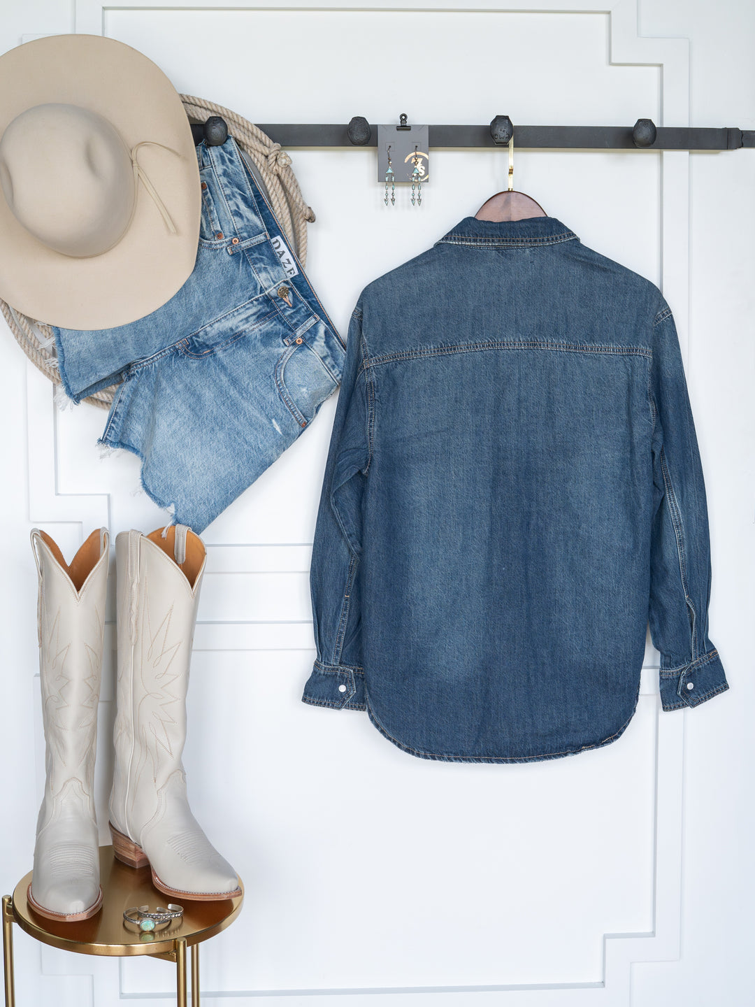 The Sorento cowboy hat by Savannah Sevens western life{&}style hangs on a wall rack with denim shorts and a distressed western yoke shirt. Below, white cowboy boots sit on a small round table.