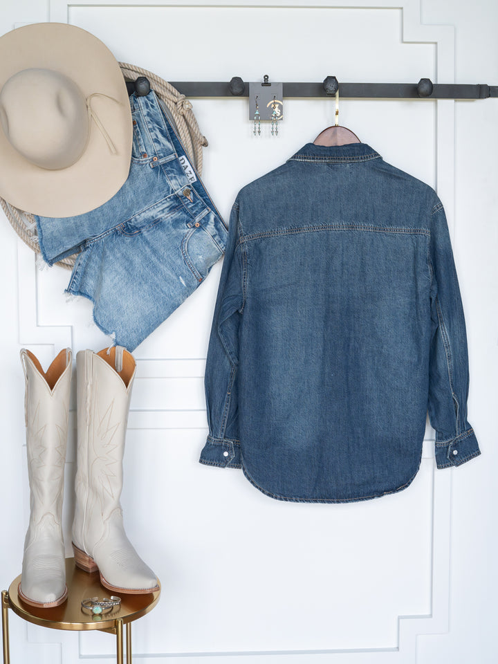 The Sorento cowboy hat by Savannah Sevens western life{&}style hangs on a wall rack with denim shorts and a distressed western yoke shirt. Below, white cowboy boots sit on a small round table.