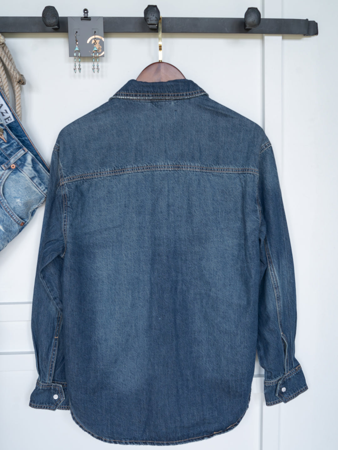 The Sorento distressed denim shirt by Savannah Sevens western life{&}style hangs on a wooden hanger against a white wall, with blue jeans and dangling earrings on a nearby hook.