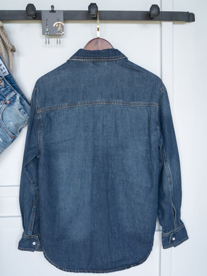 The Sorento distressed denim shirt by Savannah Sevens western life{&}style hangs on a wooden hanger against a white wall, with blue jeans and dangling earrings on a nearby hook.
