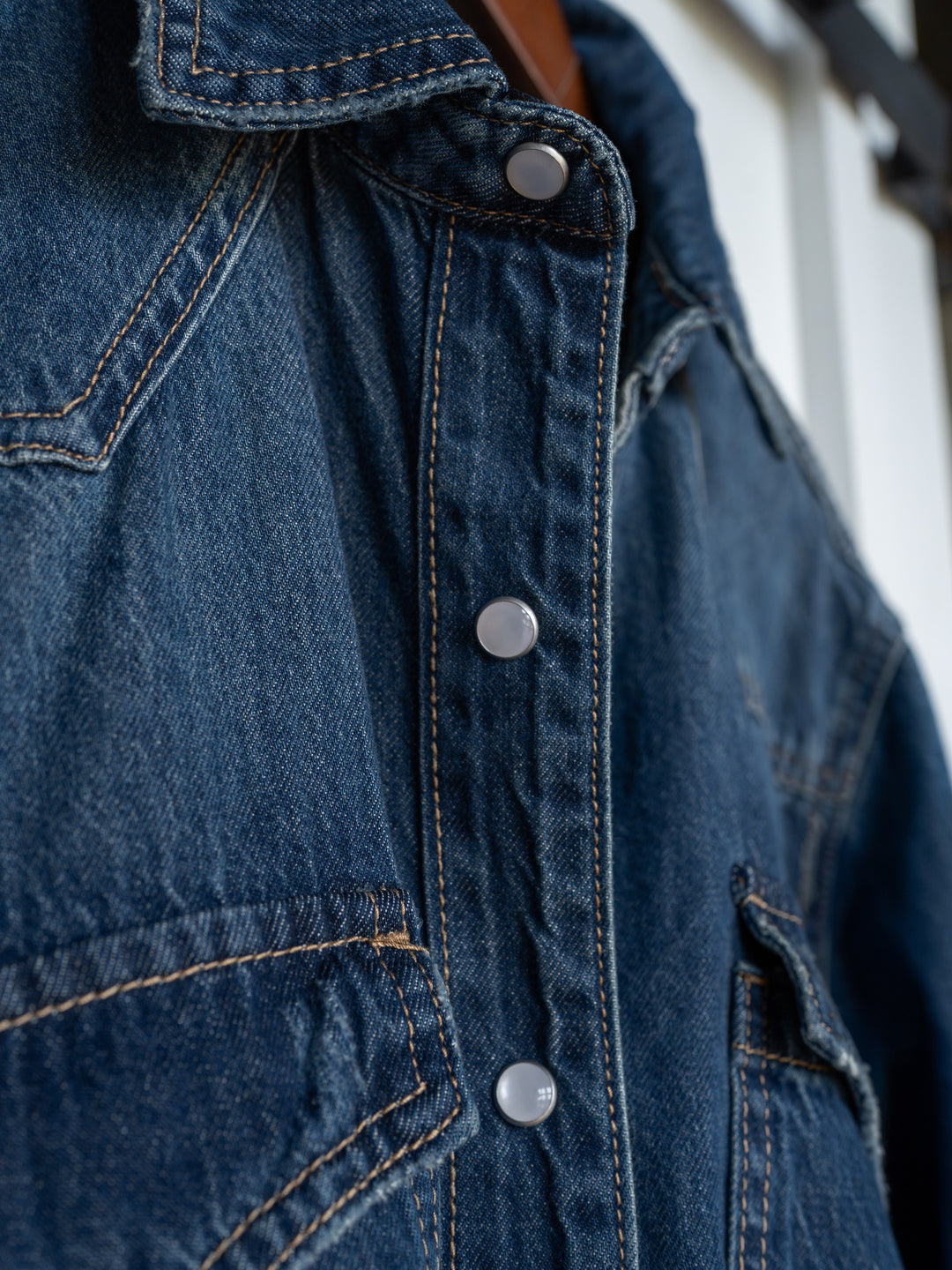 Close-up of The Sorento denim shirt by Savannah Sevens western life{&}style, featuring blue distressed fabric, western yoke, visible stitching, detailed pockets, and silver snap buttons along the front. Texture and craftsmanship are highlighted.