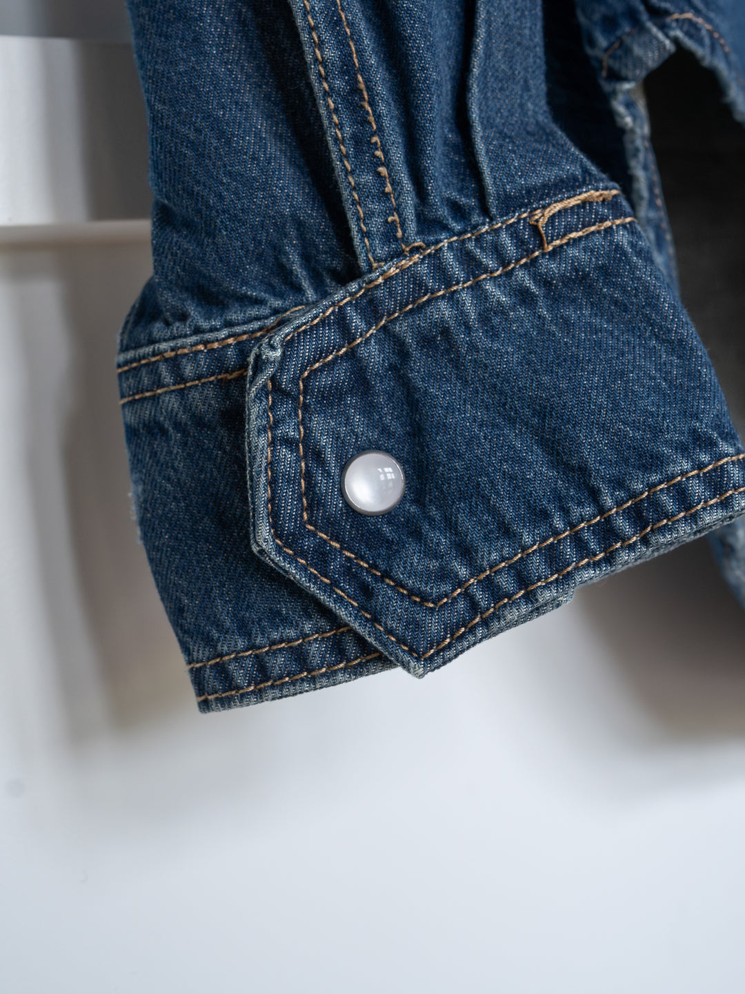 Close-up of The Sorento by Savannah Sevens western life{&}style: blue denim pearl snap top sleeve, visible orange stitching, western yoke details, and a white snap button on the cuff.