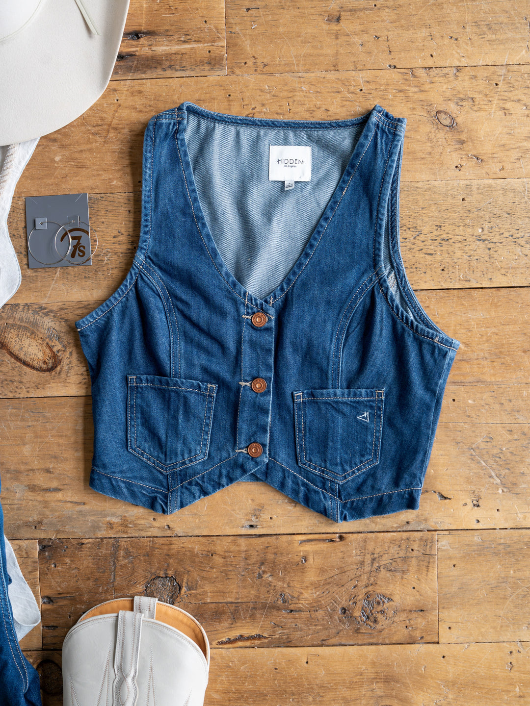 The Osage by Savannah Sevens western life{&}style, a sleeveless medium dark wash denim crop vest with three brown buttons and two front pockets, is displayed flat on wood beside a white cowboy hat, gray "7.5" tag, and white boot.