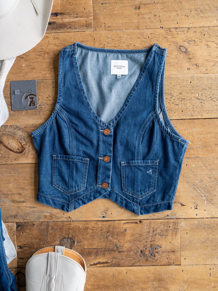 The Osage by Savannah Sevens western life{&}style, a sleeveless medium dark wash denim crop vest with three brown buttons and two front pockets, is displayed flat on wood beside a white cowboy hat, gray "7.5" tag, and white boot.