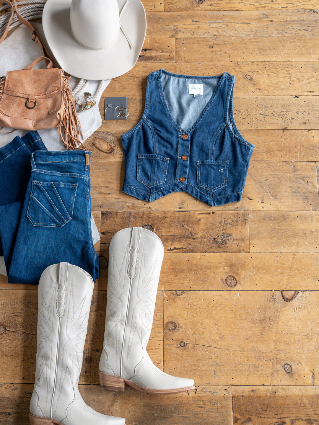 Flat lay on wooden floor featuring The Osage white cowboy boots from Savannah Sevens western life{&}style, medium dark wash jeans, a denim crop vest, white cowboy hat, tan fringe purse, and gray t-shirt.