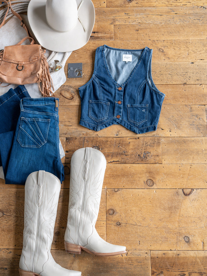 Flat lay on wooden floor featuring The Osage white cowboy boots from Savannah Sevens western life{&}style, medium dark wash jeans, a denim crop vest, white cowboy hat, tan fringe purse, and gray t-shirt.