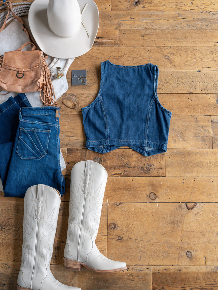 Flat lay of The Osage by Savannah Sevens western life{&}style: white cowboy hat, tan fringe purse, denim crop vest (medium dark wash), blue jeans, white cowboy boots, and silver jewelry on wooden floor.
