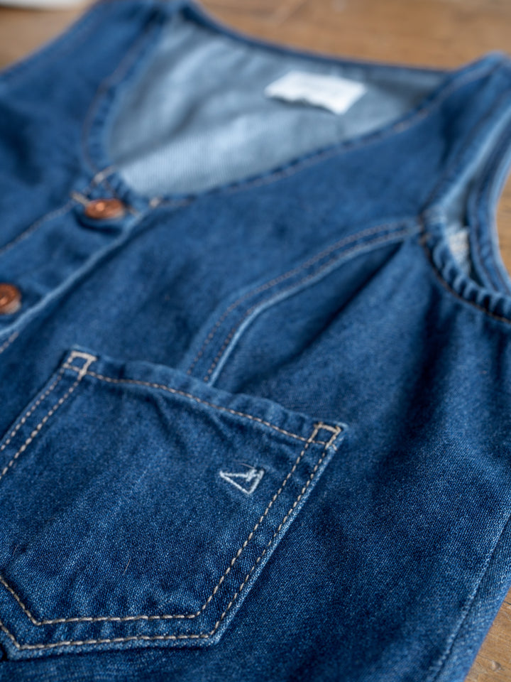 The Osage by Savannah Sevens western life{&}style is a medium dark wash denim crop vest with brown buttons, front pocket, and visible stitching, shown laid flat on wood.