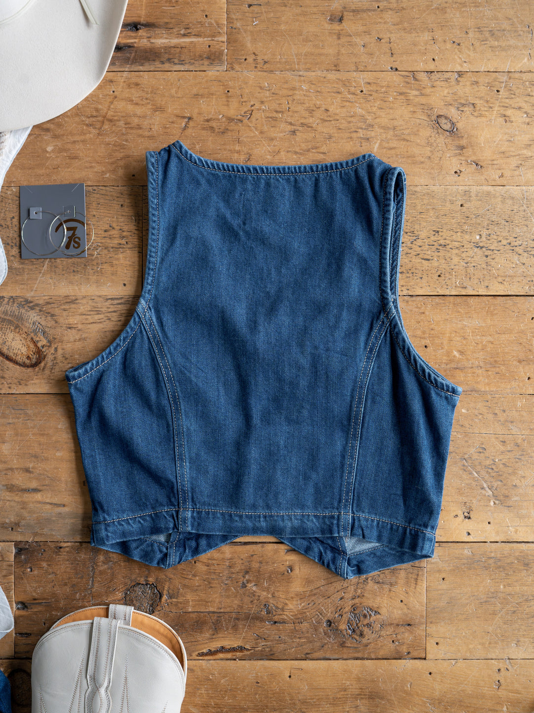 The Osage by Savannah Sevens western life{&}style, a medium dark wash denim crop vest, is shown laid flat on wood with its back visible. A white hat, white handbag, and a gray box marked "7" are partially seen at the edges.