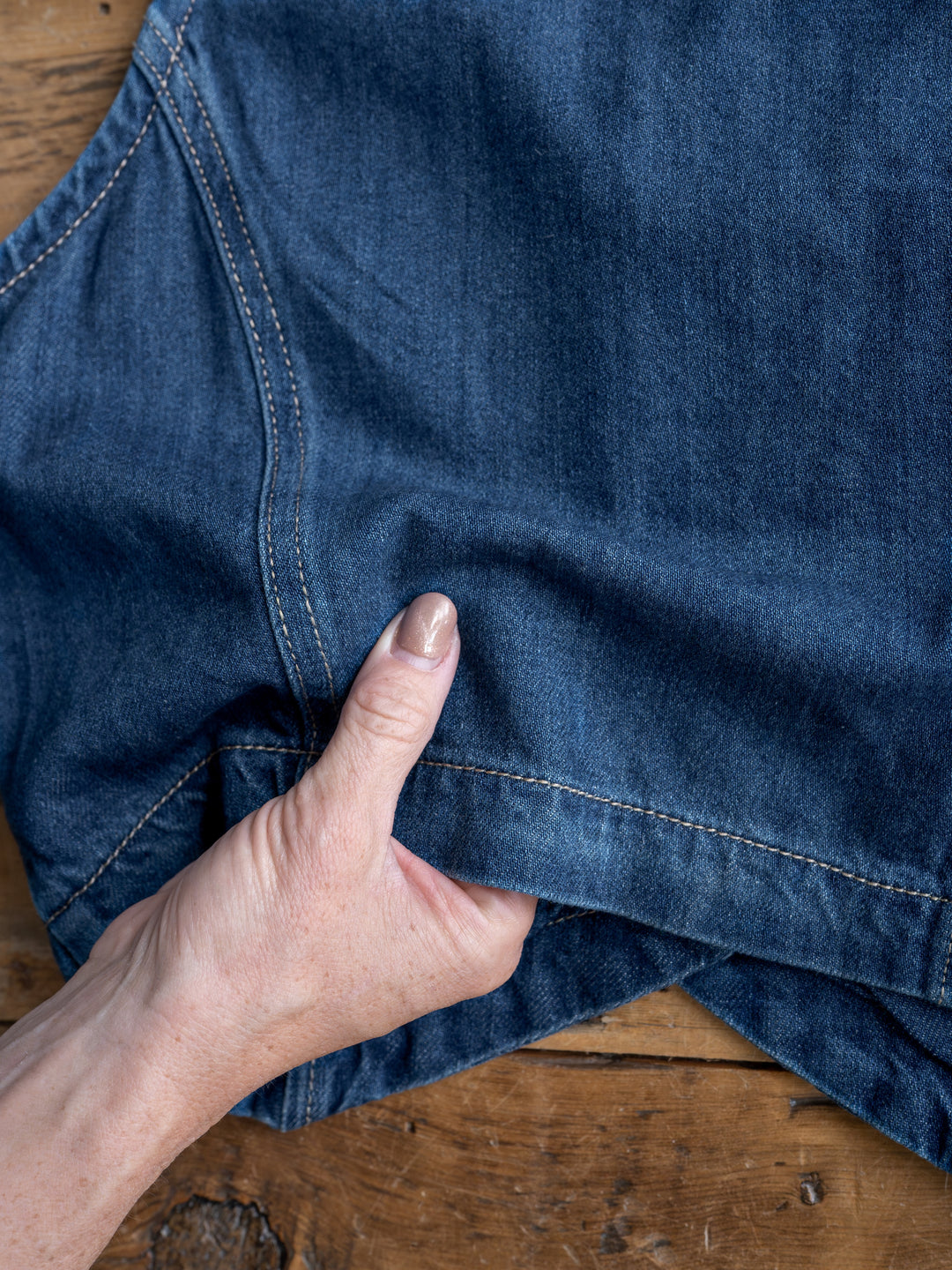 A hand pinches the hem of The Osage medium dark wash blue denim jeans by Savannah Sevens western life{&}style, shown on a wooden surface.