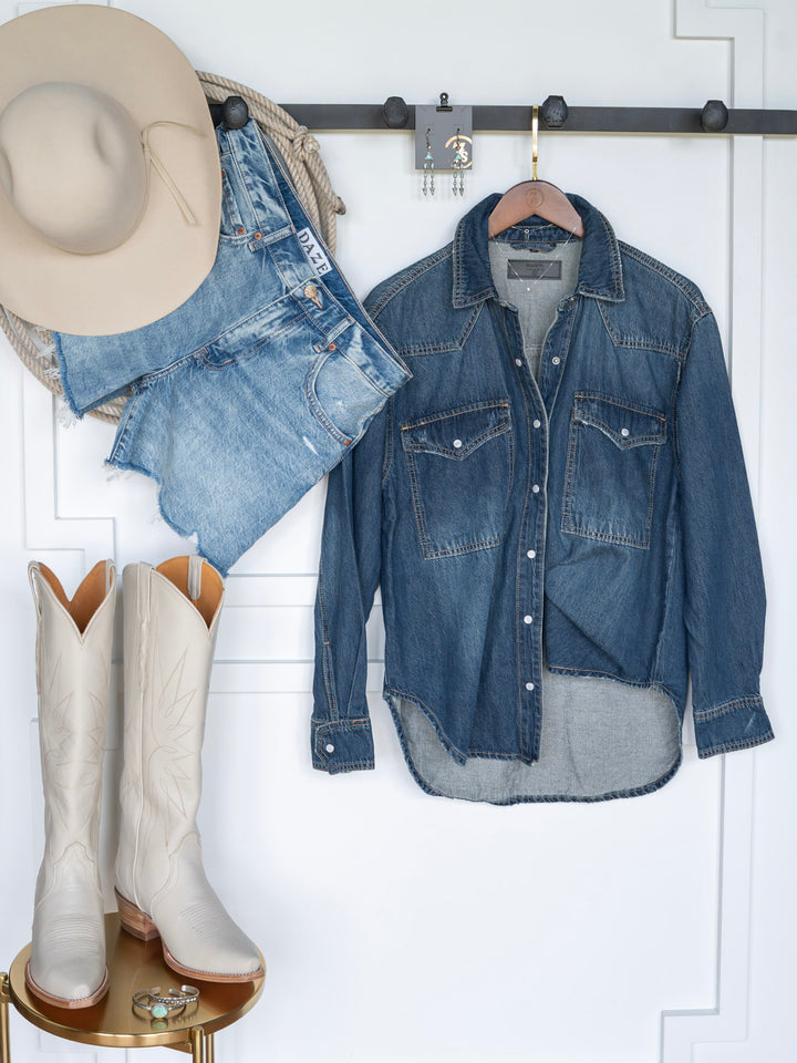 The Sorento by Savannah Sevens western life{&}style: a beige cowboy hat, distressed denim shorts, denim pearl snap top on a hanger, and white cowboy boots displayed against a white wall.