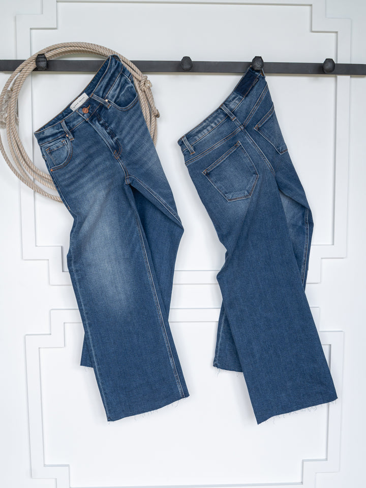 Two pairs of The Britton High Rise Wide Leg Crop Jeans by Savannah Sevens western life{&}style hang on a black rack against a white wall, accented with a coiled rope. These jeans offer classic front and back pockets for timeless style.