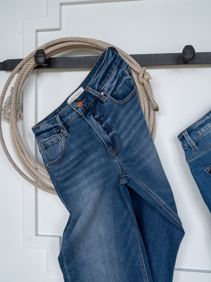 The Britton High Rise Wide Leg Crop Jean by Savannah Sevens western life{&}style hangs on a black metal hook with a coiled rope against a white wall.