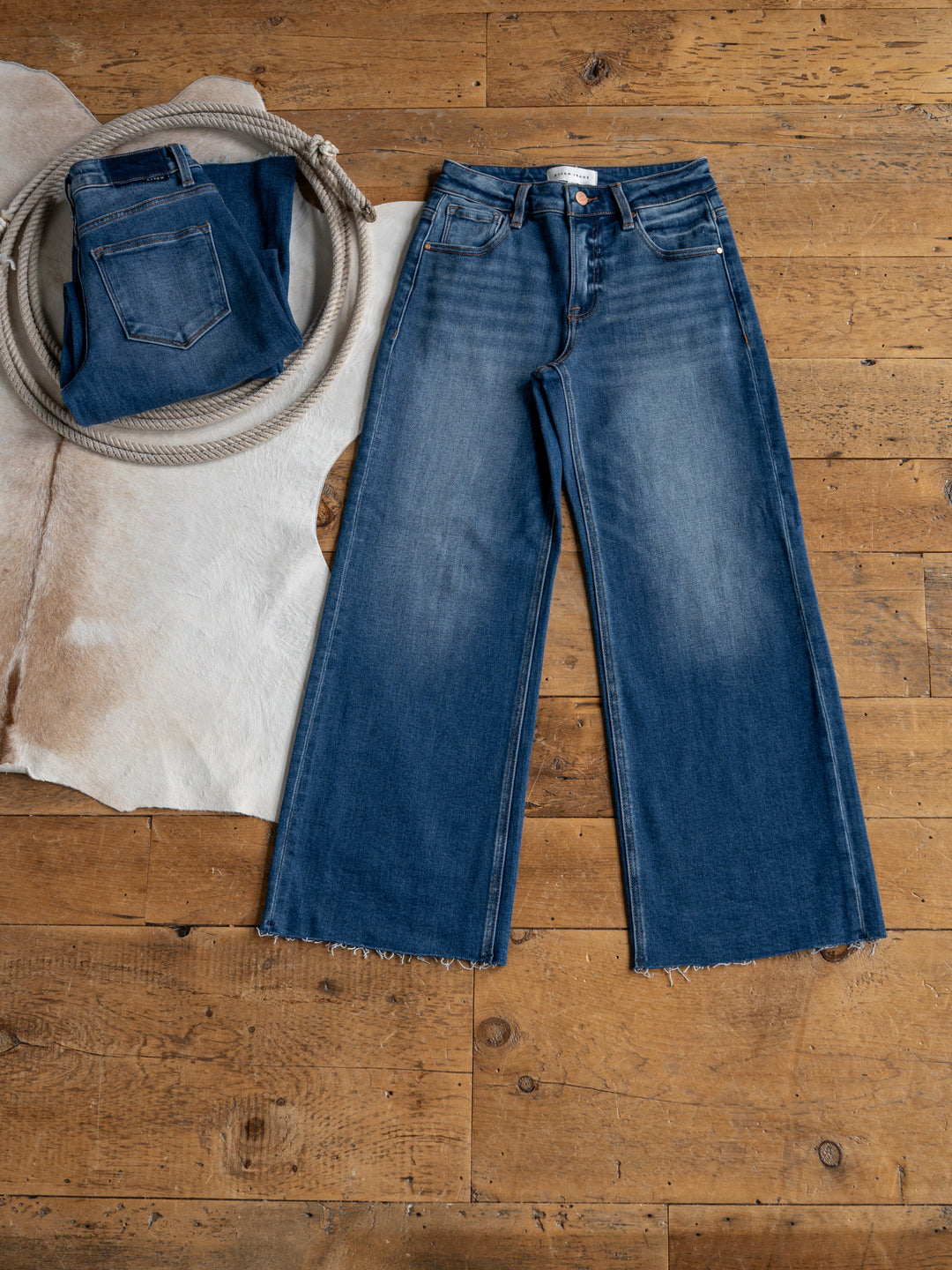 The Britton High Rise Wide Leg Crop Jean by Savannah Sevens western life{&}style lies flat on a wooden floor next to a folded jean, coiled rope, and cowhide rug.