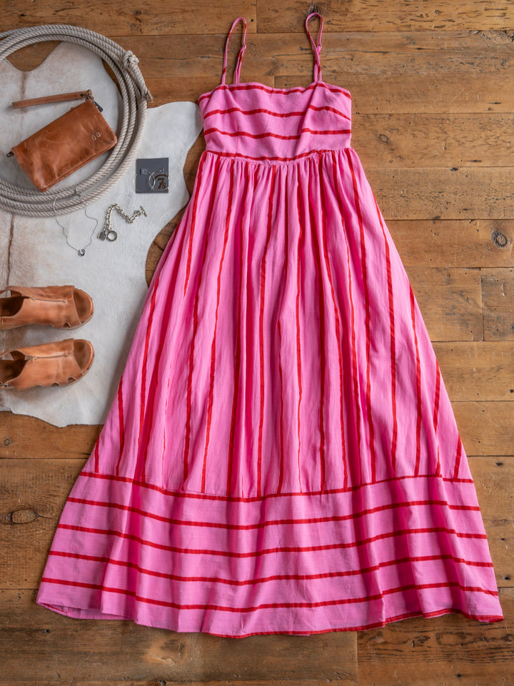 The Fredonia dress by Savannah Sevens western life{&}style, featuring pink and red stripes and a sleeveless design, is styled on a wooden floor with tan sandals, a brown clutch bag, and a necklace placed nearby.