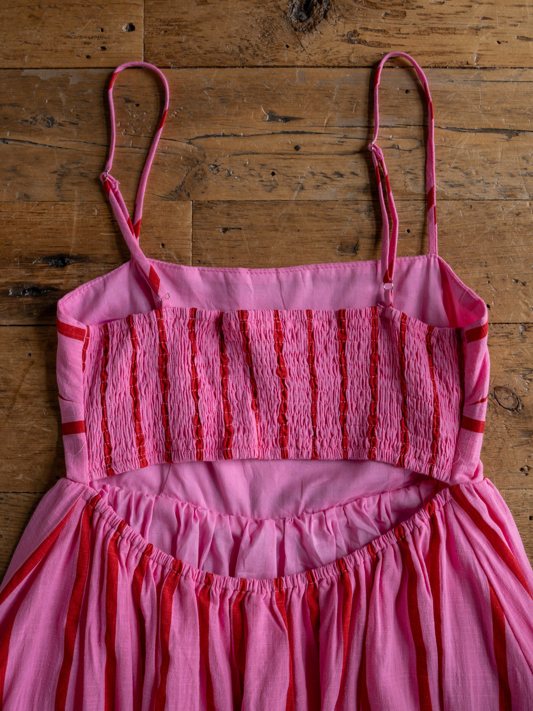 A close-up of The Fredonia by Savannah Sevens western life{&}style, a pink and red striped sleeveless dress with spaghetti straps and smocked bodice, laid flat on a wooden surface.