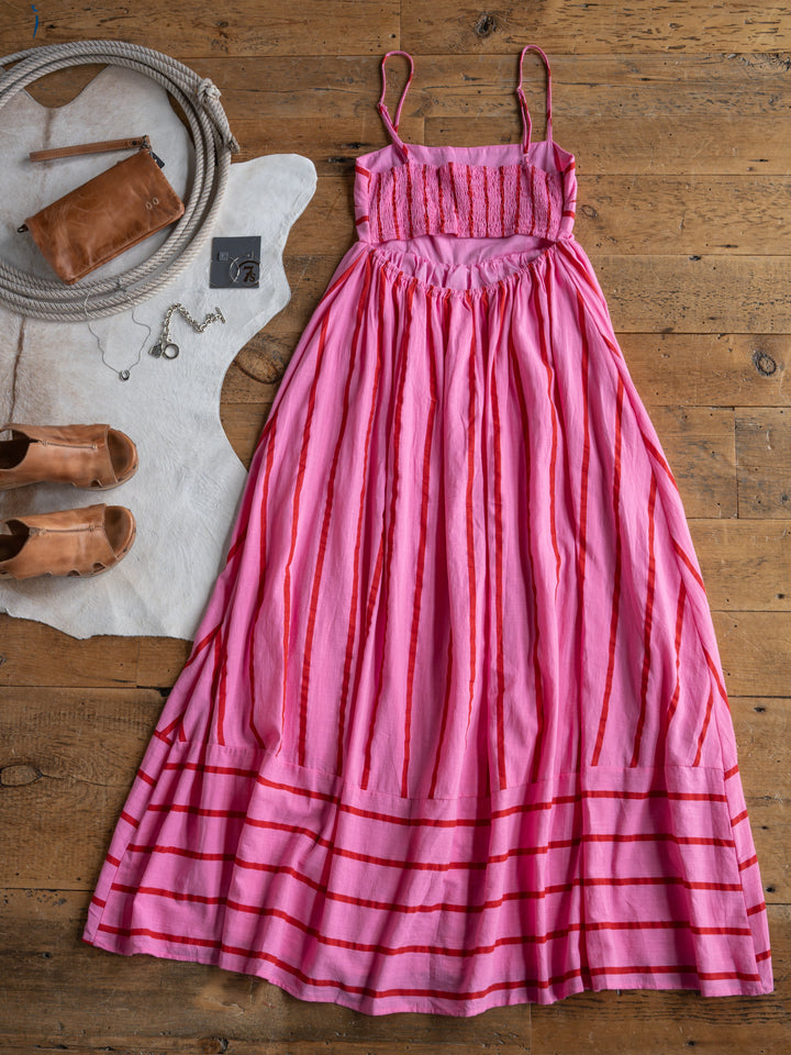 The Fredonia dress by Savannah Sevens western life{&}style, in pink and red stripes, is displayed on a wooden floor with tan sandals, a brown clutch, a necklace, and a white hide rug.