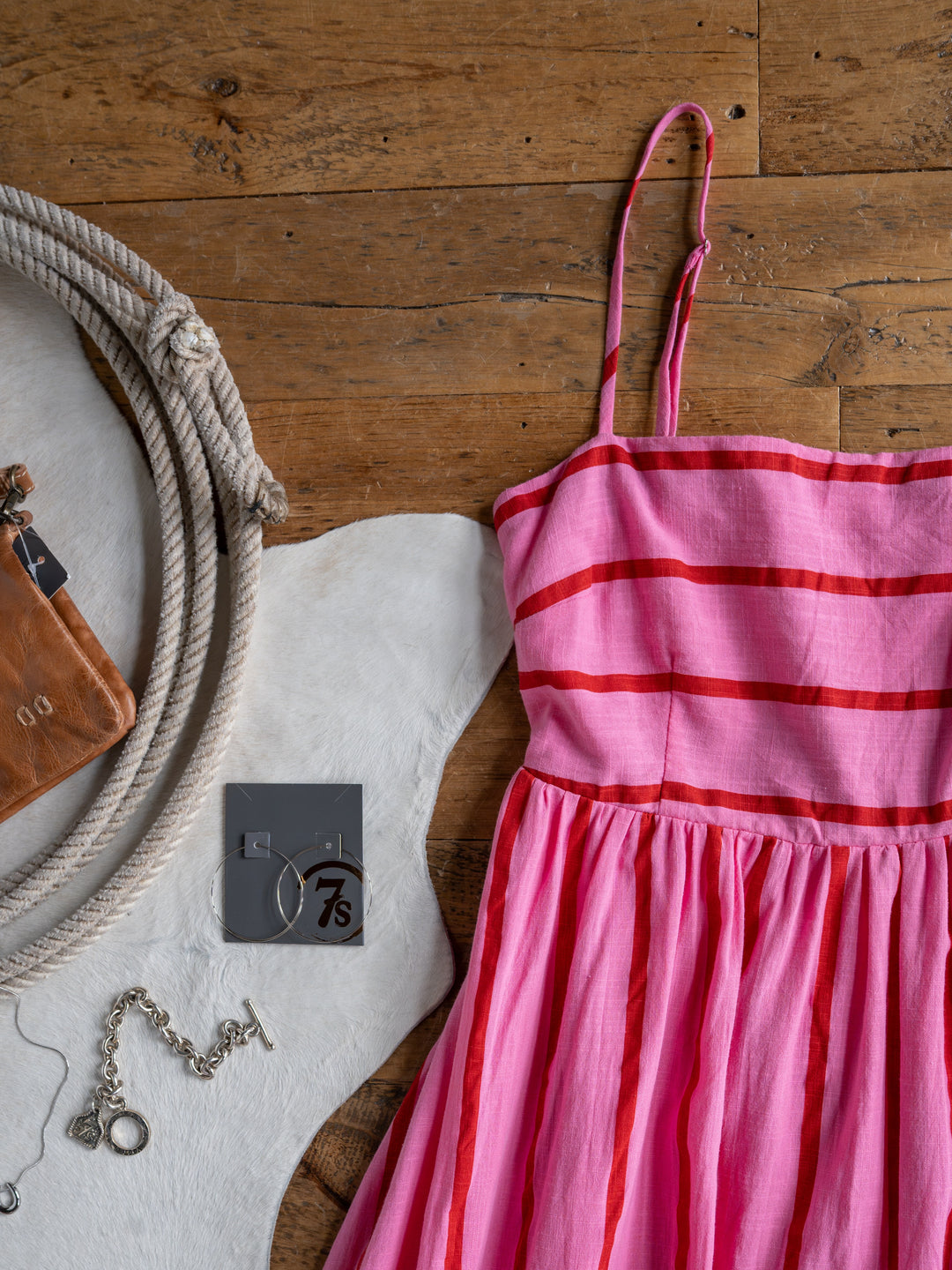 The Fredonia sleeveless pink and red striped dress by Savannah Sevens western life{&}style is displayed on wood with a silver bracelet, hoop earrings, and a small brown leather pouch.