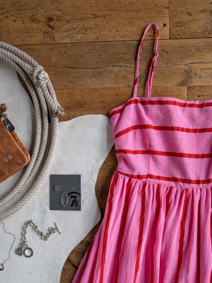 The Fredonia sleeveless pink and red striped dress by Savannah Sevens western life{&}style is displayed on wood with a silver bracelet, hoop earrings, and a small brown leather pouch.