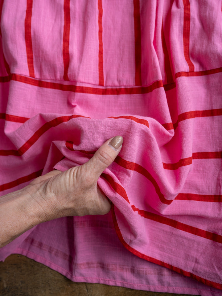 A hand gently scrunches the bold striped pink and red fabric of The Fredonia sleeveless dress by Savannah Sevens western life{&}style, highlighting its texture and drape.