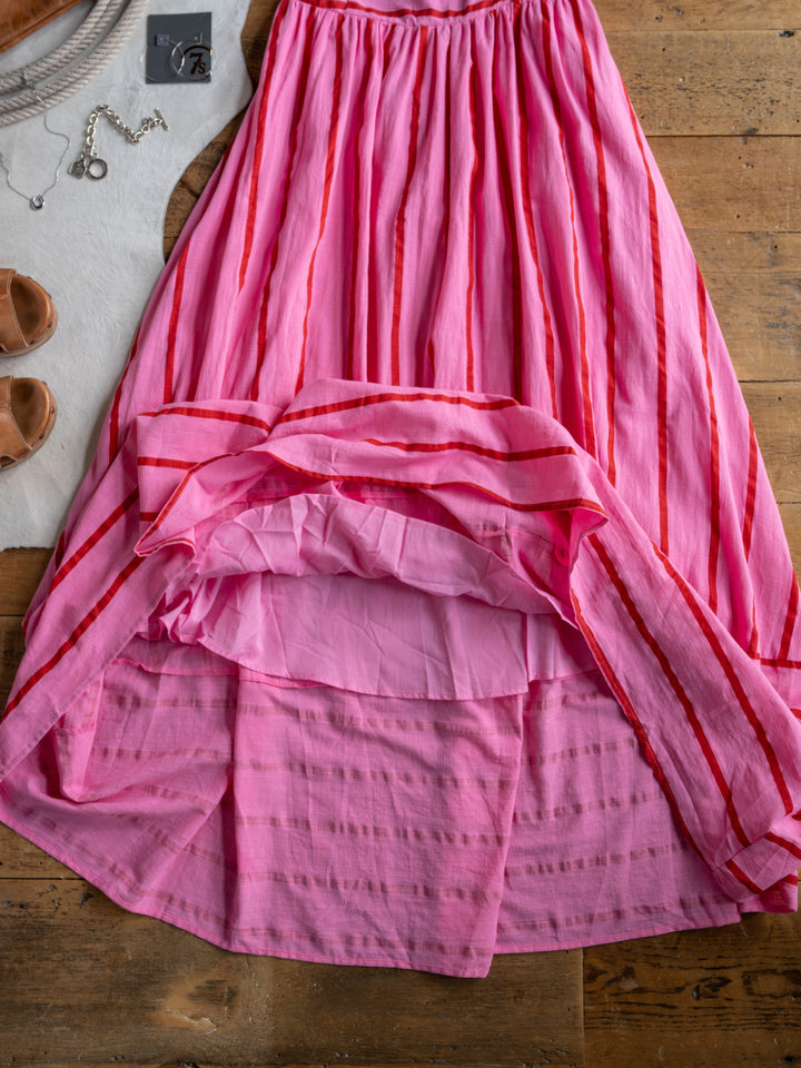The Fredonia by Savannah Sevens western life{&}style, a pink and red striped maxi skirt, is displayed on a wooden floor with tan sandals, silver jewelry, and a white accent mat.
