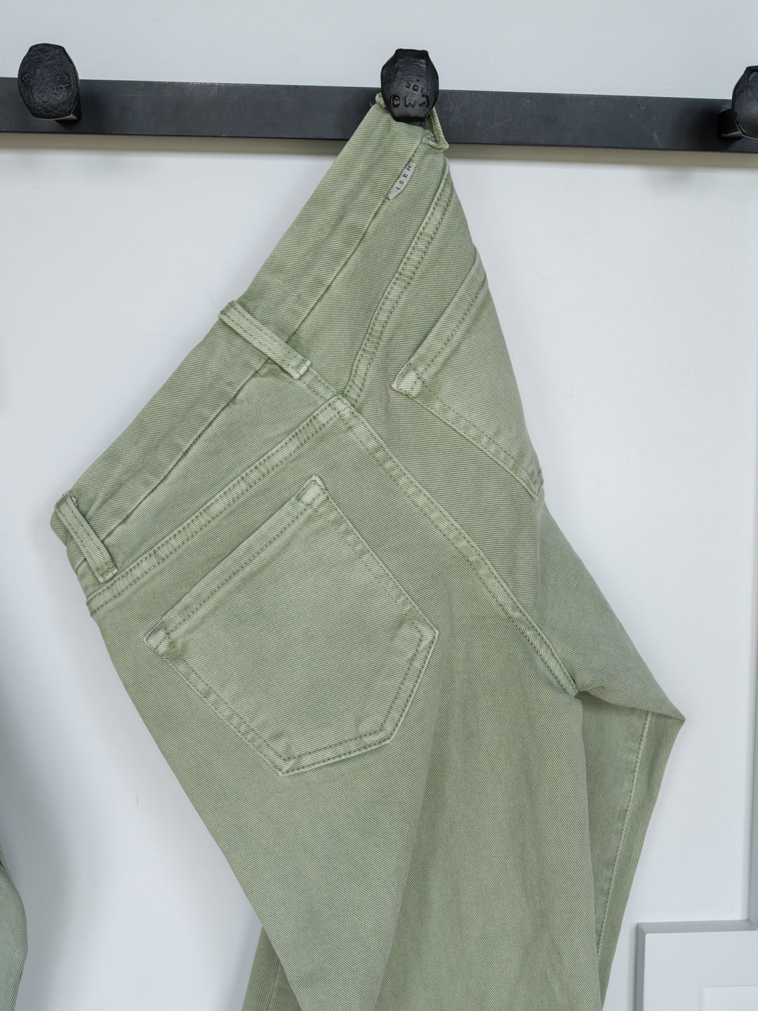The Lindy High Rise Wide Leg Jean by Savannah Sevens western life{&}style hangs by a belt loop on a black hook against a white background, displaying the sage denim’s back pockets, seams, and raw cut hem.