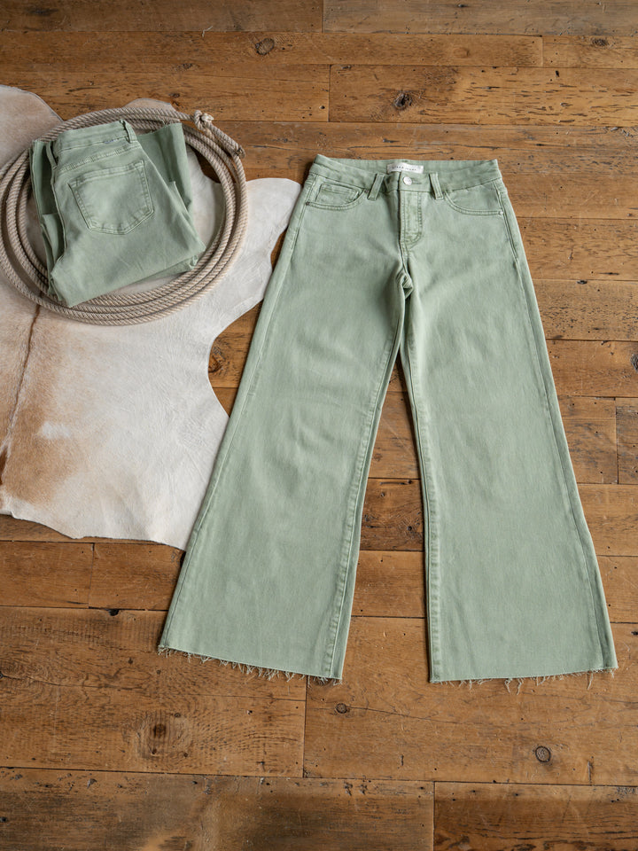 The Lindy High Rise Wide Leg Jean by Savannah Sevens western life{&}style is laid flat on a wooden floor beside a folded pair, coiled rope, and tan cowhide rug, featuring stylish frayed hems for an effortlessly cool look.