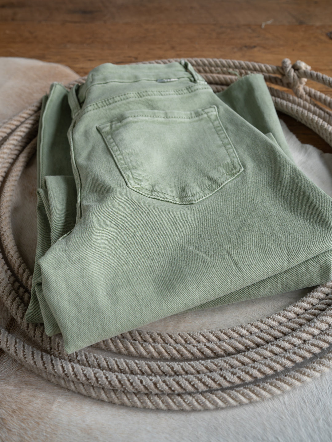The Lindy High Rise Wide Leg Jean by Savannah Sevens western life{&}style in sage denim, featuring a raw cut hem, is neatly folded on a light cowhide rug with a coiled rope nearby on a wooden floor.