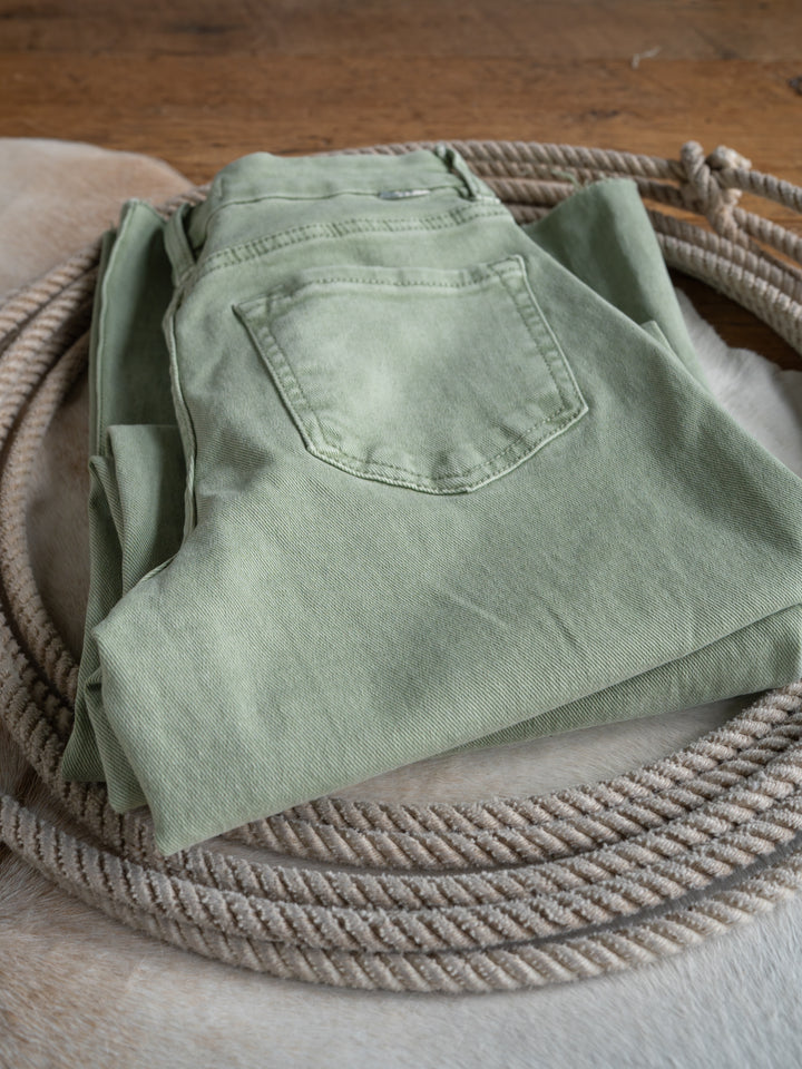 The Lindy High Rise Wide Leg Jean by Savannah Sevens western life{&}style in sage denim, featuring a raw cut hem, is neatly folded on a light cowhide rug with a coiled rope nearby on a wooden floor.