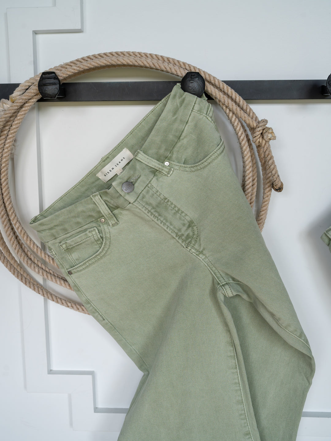 The Lindy High Rise Wide Leg Jean by Savannah Sevens western life{&}style in sage denim hangs on a black rack, featuring a raw cut hem, with a coiled rope behind it against a white paneled background.
