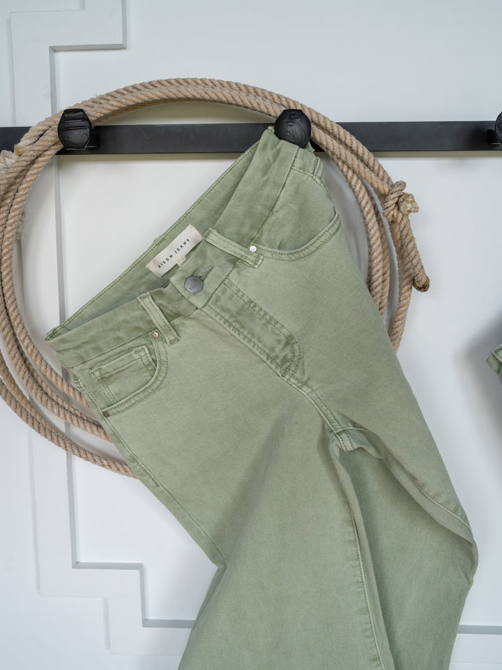 The Lindy High Rise Wide Leg Jean by Savannah Sevens western life{&}style in sage denim hangs on a black rack, featuring a raw cut hem, with a coiled rope behind it against a white paneled background.
