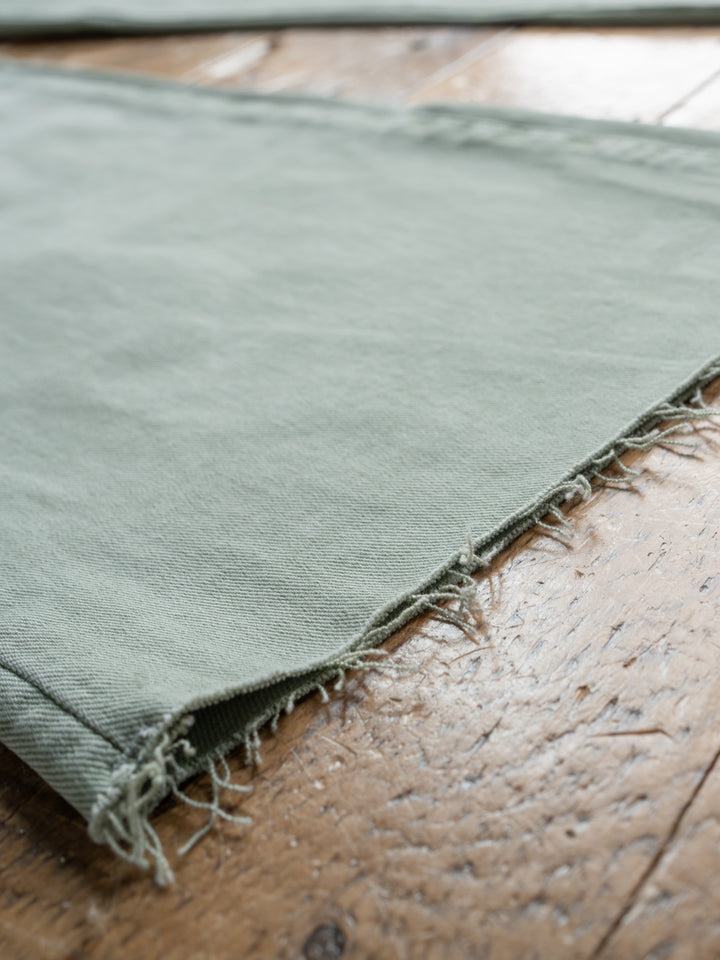 Close-up of light green fabric with a raw cut hem, lying on wood, showing texture—reminiscent of The Lindy High Rise Wide Leg Jean by Savannah Sevens western life{&}style.