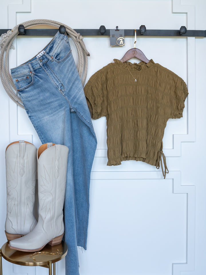 The Talmo jeans by Savannah Sevens western life{&}style, shown with a ruched olive short sleeve top and white cowboy boots, are displayed against a white wall with decorative trim.
