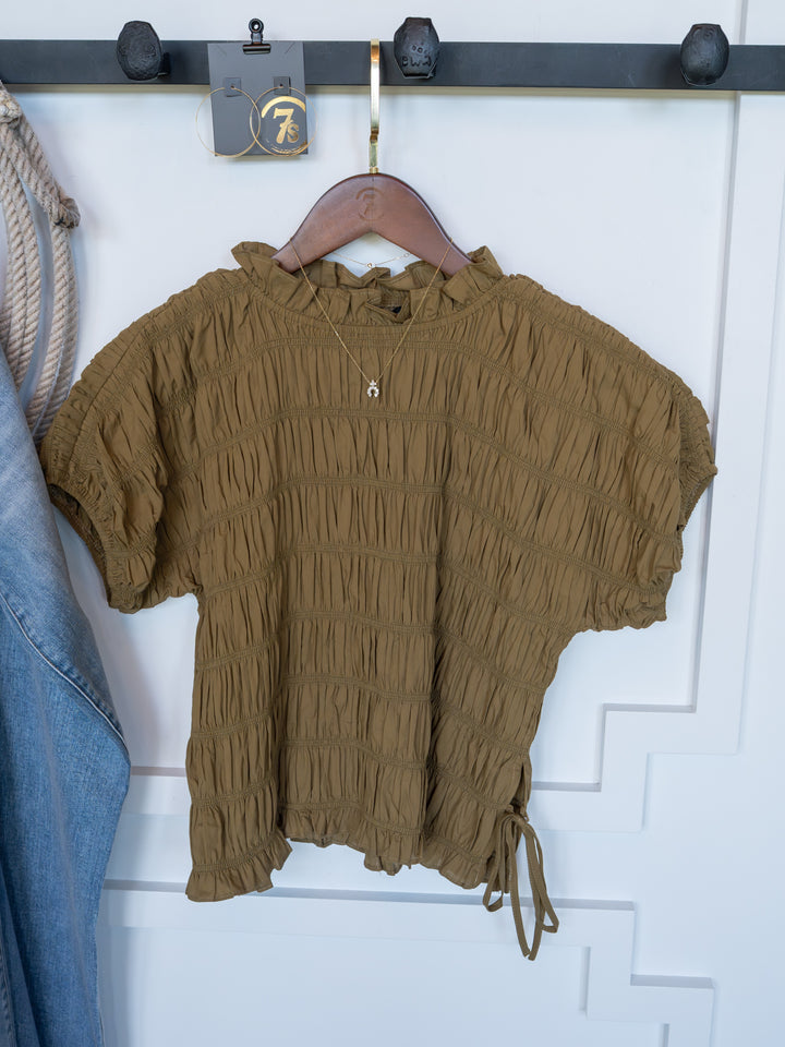 The Talmo by Savannah Sevens western life{&}style, a brown ruched blouse with short sleeves, hangs on a wooden hanger against a white wall with a gold necklace. A blue denim piece is partially visible on the left.