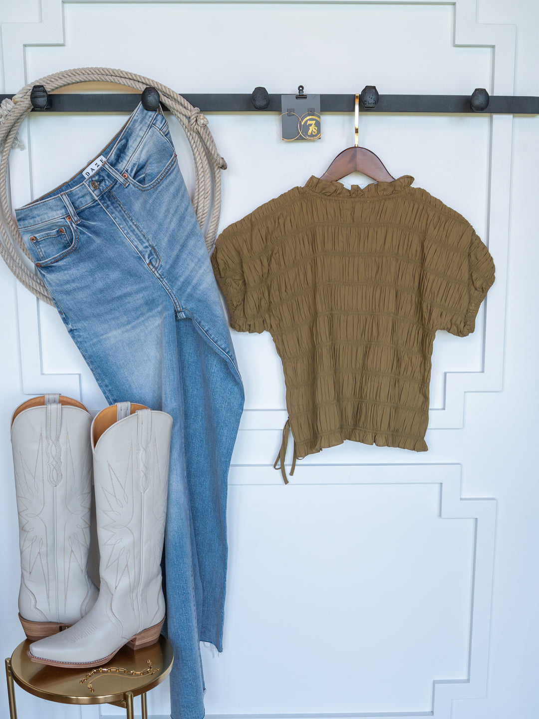 The Talmo jeans and an olive ruffle-neck short sleeve top by Savannah Sevens western life{&}style hang on hooks, while tall white cowboy boots sit on a round table against a white wall.