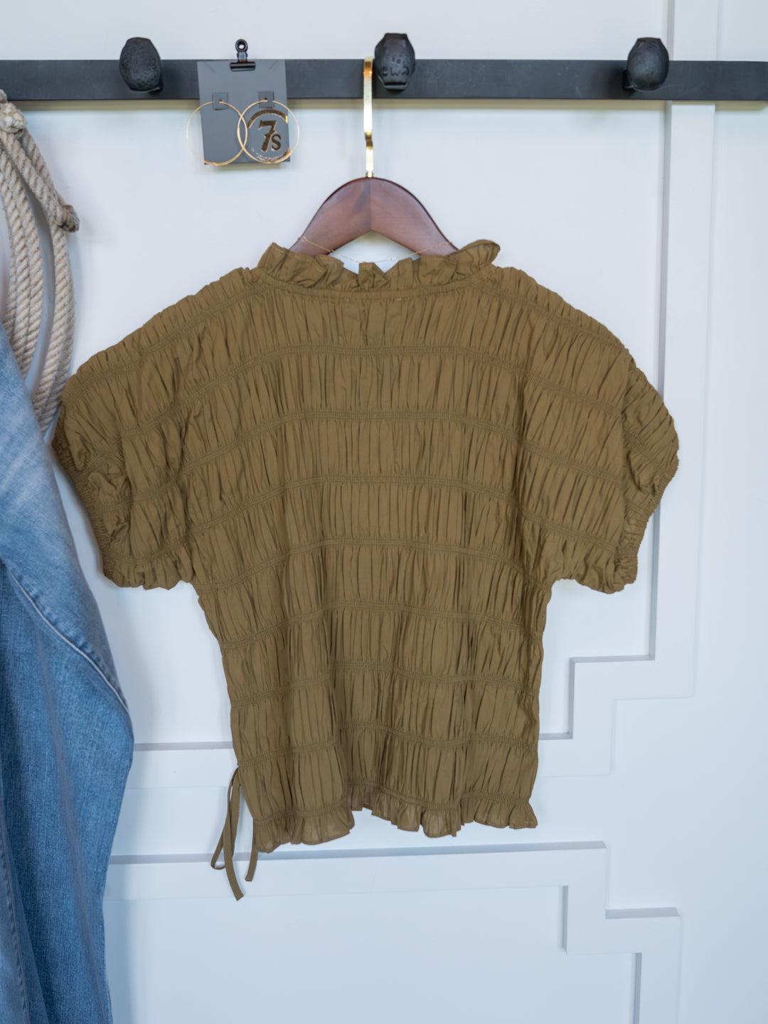 The Talmo by Savannah Sevens western life{&}style is a brown short-sleeve blouse with ruched design and ruffle neckline, displayed on a wooden hanger beside denim and thick rope against a white paneled wall.