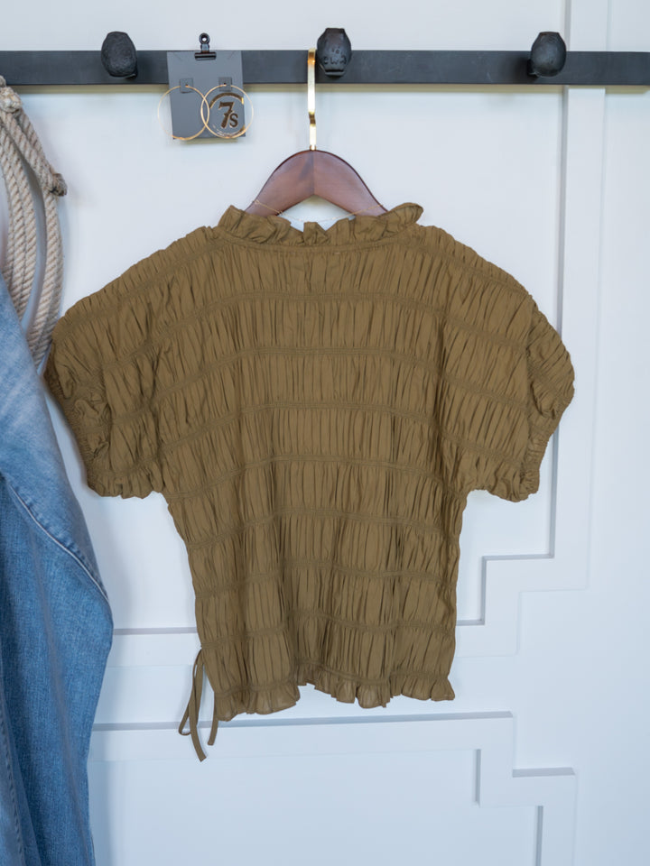 The Talmo by Savannah Sevens western life{&}style is a brown short-sleeve blouse with ruched design and ruffle neckline, displayed on a wooden hanger beside denim and thick rope against a white paneled wall.