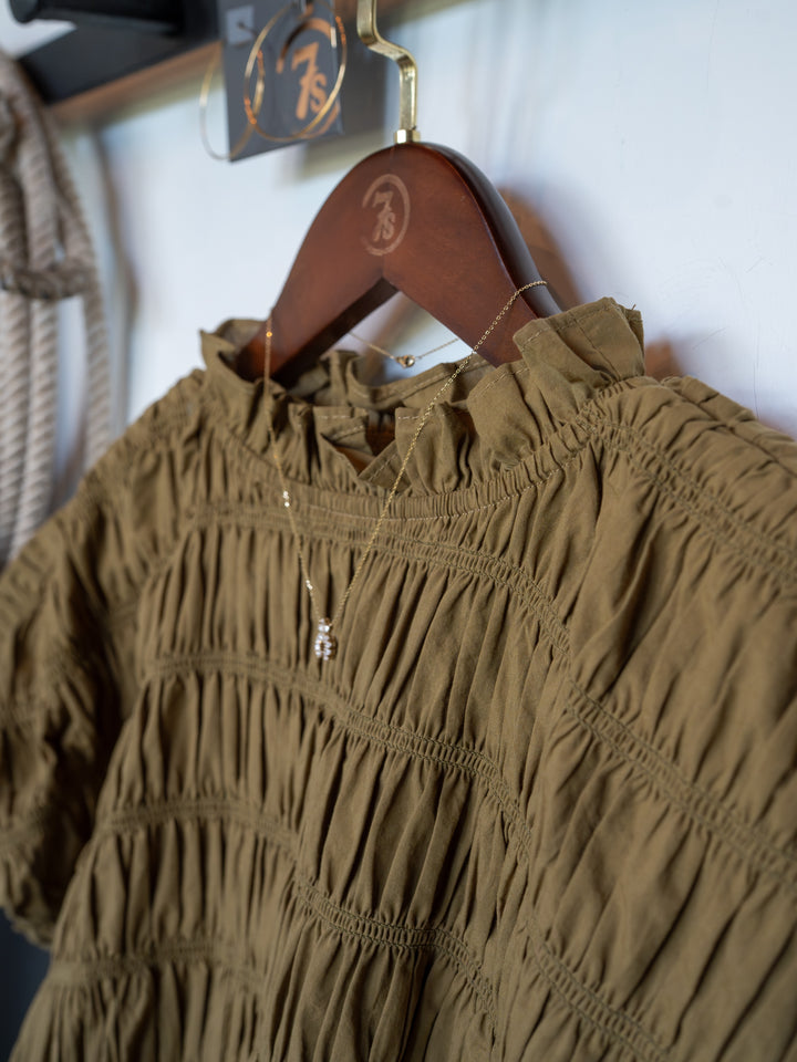 A close-up of The Talmo brown blouse by Savannah Sevens western life{&}style, featuring a ruched design and ruffle neckline on a wooden hanger with a gold necklace. The background shows part of a rope and a clear sign with gold writing.