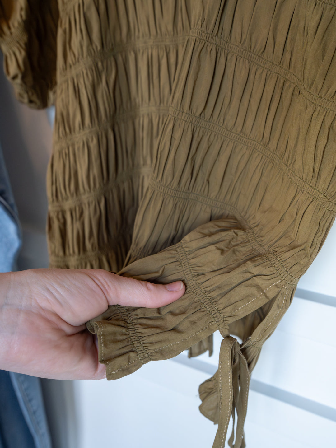 A hand holds the sleeve of The Talmo blouse by Savannah Sevens western life{&}style, a light brown, crinkled-texture top with ruched details, tie cuff, gathered seams, and a ruffle neckline, shown against a white background.