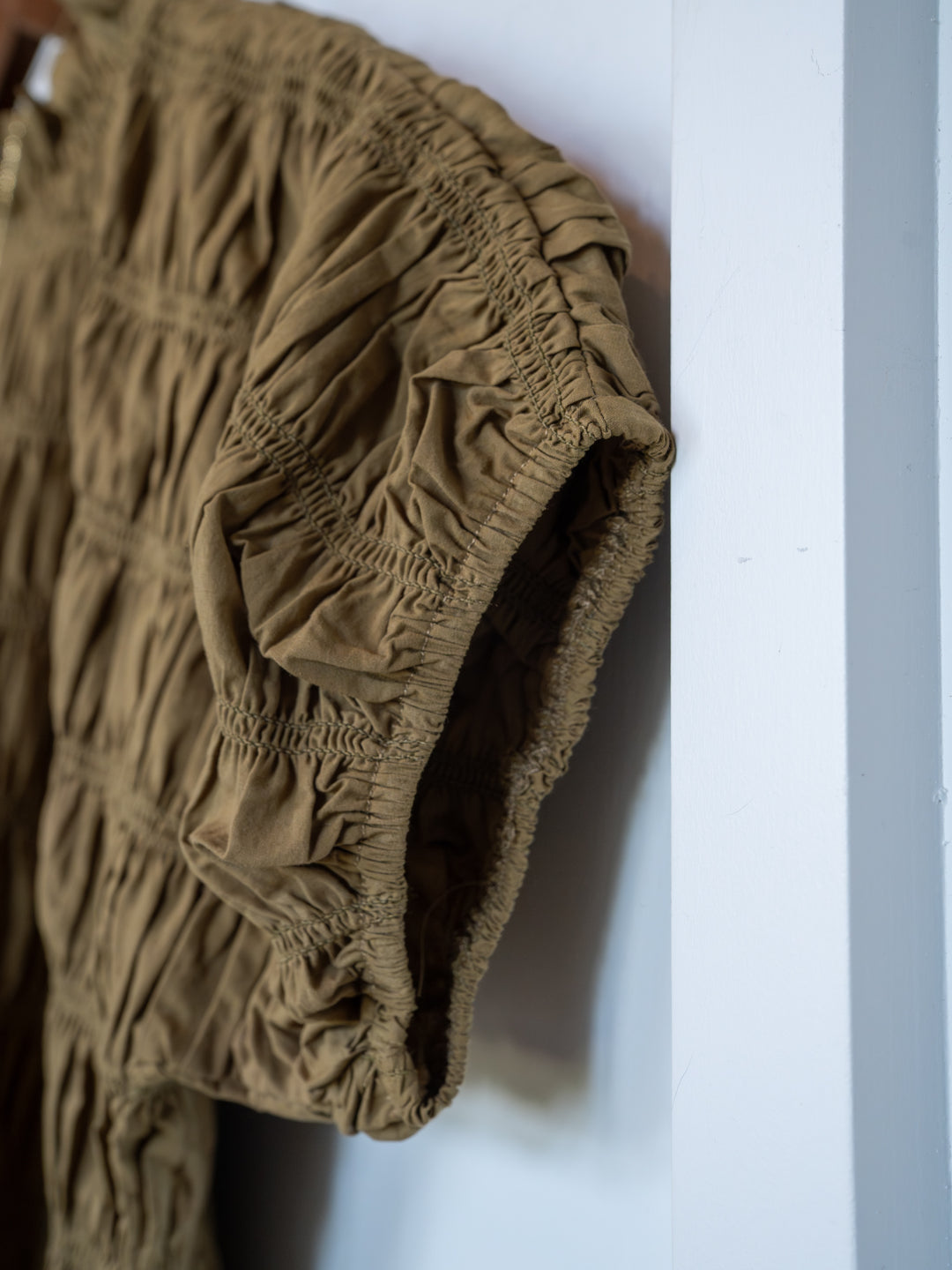 Close-up of The Talmo olive short sleeve top by Savannah Sevens western life{&}style, featuring a ruched design with gathered fabric texture and elastic detailing, hanging against a white wall.