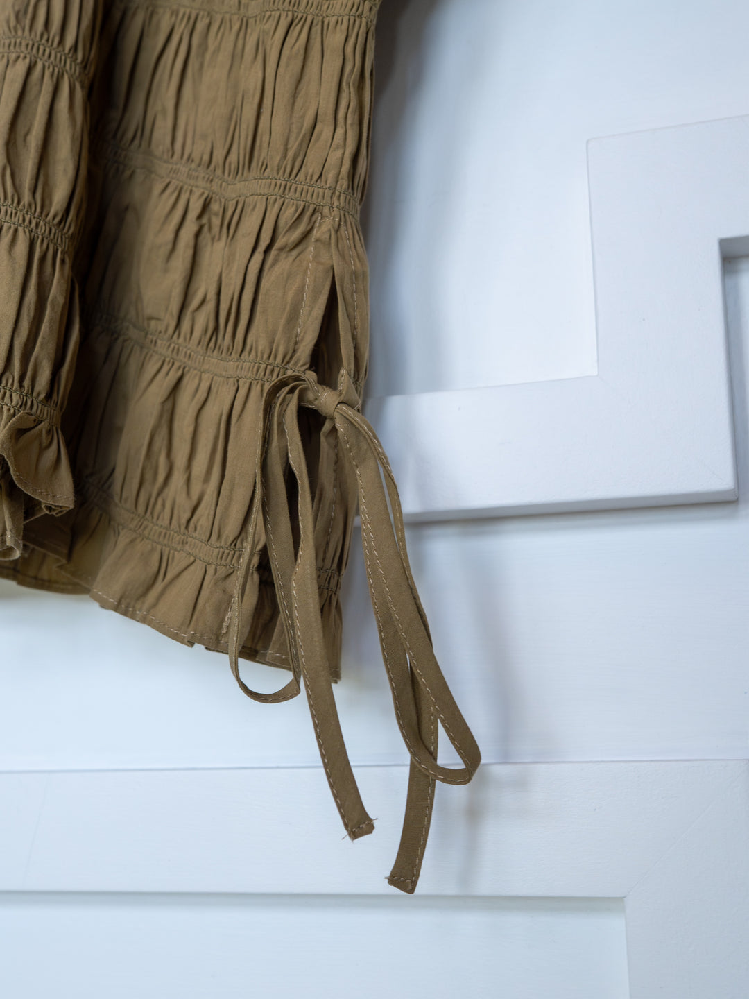 A close-up of The Talmo by Savannah Sevens western life{&}style, a brown ruched garment with tie detail and ruffle neckline, hanging against a white geometric-patterned background.