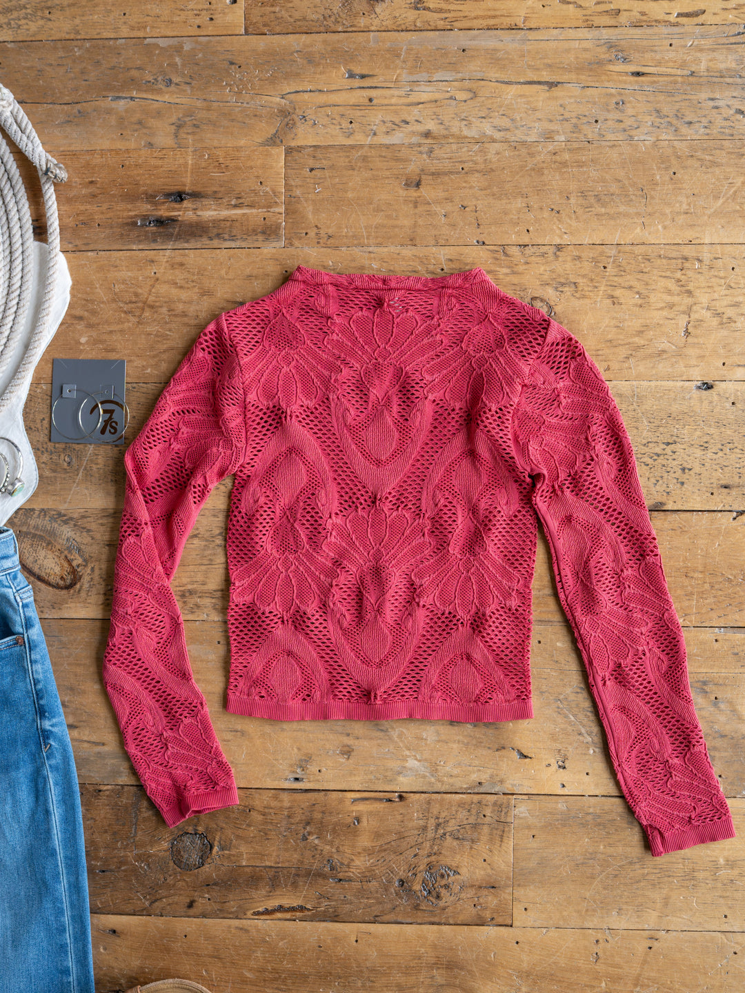 The Lexa by Savannah Sevens western life{&}style, a bright pink long sleeve sheer crochet floral top, is laid flat on a wooden floor beside blue jeans, a gray tag, and a white coiled rope.