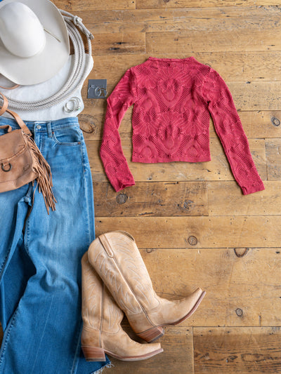 Flat lay of The Lexa, a raspberry red long sleeve crochet top by Savannah Sevens western life{&}style, styled with a white hat, ivory rope belt, brown fringe purse, blue jeans, tan cowboy boots, and silver belt buckle on a wooden floor.